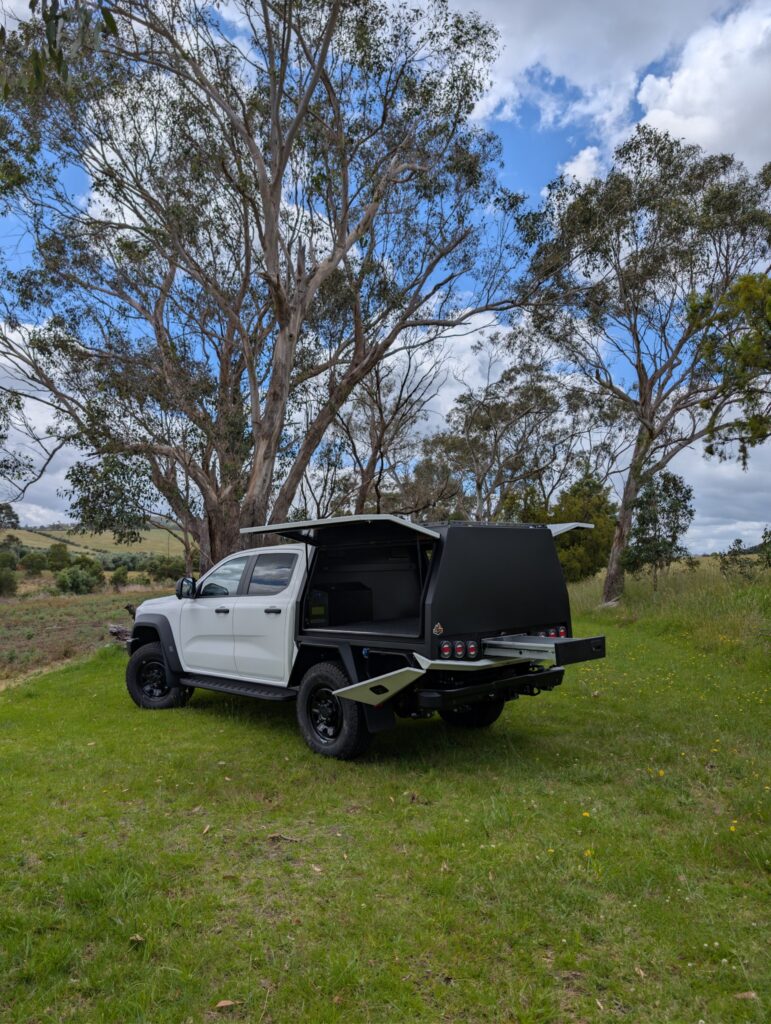 White ute with black Trig Point chassis-mount canopy, gullwing door open, rear trundle drawer extended, parked in a paddock
